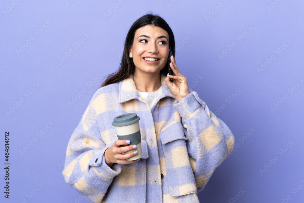 Young brunette woman over isolated purple background holding coffee to take away and a mobile Young brunette woman over isolated purple background holding coffee to take away and a mobile