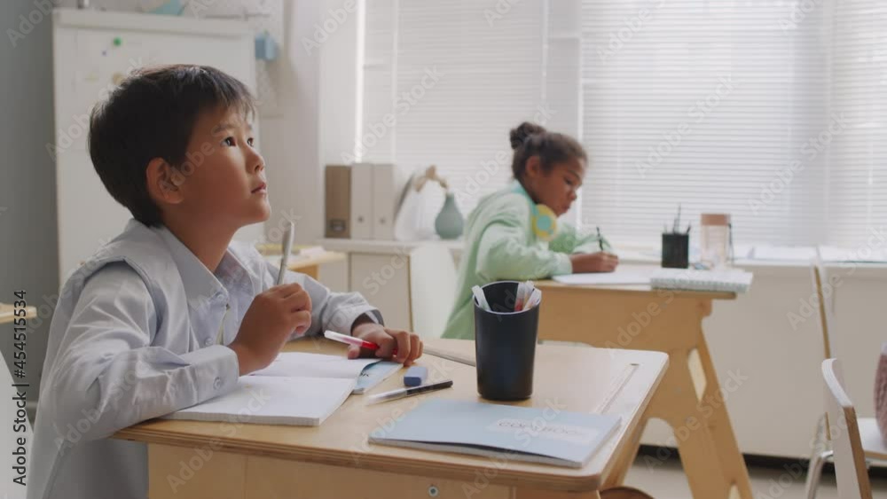 Handheld tracking shot of 12-year-old Asian boy sitting at desk in classroom and taking test or solving math equation Handheld tracking shot of 12-year-old Asian boy sitting at desk in classroom and taking test or solving math equation