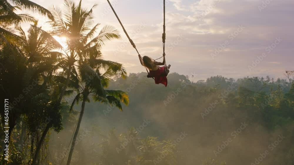 travel woman swinging over jungle at sunrise enjoying exotic vacation sitting on swing with sun flare shining through palm trees in tropical rainforest holiday lifestyle freedom