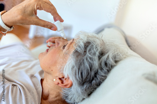 Side view of senior gray hair woman applying eye drop at home. Senior woman suffering from an ophthalmologic allergic reaction to pollen. Woman applying artificial tears, antibiotics, antihistamine.