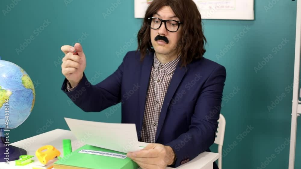 A funny school teacher with a mustache in glasses sits in the class and checks the test. A funny school teacher with a mustache in glasses sits in the class and checks the test.