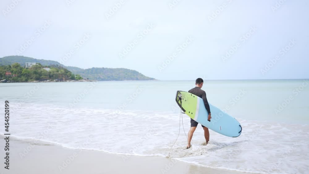 4K Confidence Asian man in wetsuit holding surfboard and walking on tropical beach at summer sunny day. Healthy sportsman enjoy outdoor active lifestyle play extreme sport surfing on summer vacation