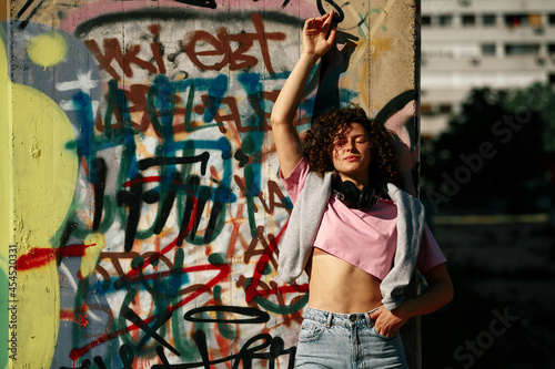 A young woman with curly hair stands against the wall and enjoying the sunny day. The woman has headphones around her neck, and on the wall, there is graffiti.