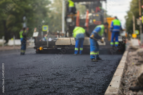 Process of asphalting, blacktopping and paving, group of workers with shovels finishing the asphalt layer, with asphalt paver machine and steam roller compactor vehicle in the background © tsuguliev