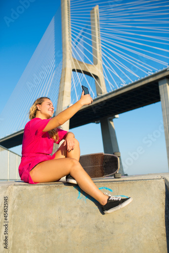 Attractive young woman taking selfie. Beautiful girl in crimson dress with skateboard sitting near bridge, taking photo of herself. Sport, hobby, active lifestyle, social media, photo concept