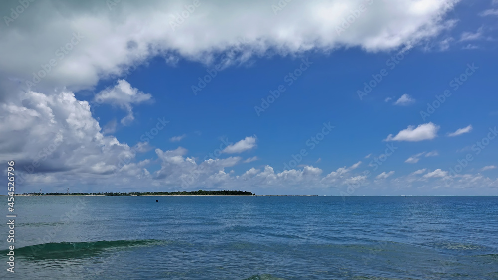There are small waves and ripples on the aquamarine ocean. There are picturesque cumulus clouds in the azure sky. A tropical island is visible in the distance. Maldives