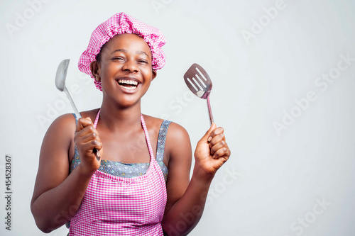 cheerful african lady holding kitchen wares, black girl in apron and a head wrap excited- indoor food concept