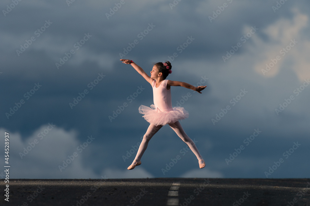 Obraz premium Little ballet dancer girl wearing a pink tutu jumping in the horizon of the road. Dramatic sky with copy space in the background
