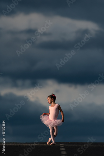 Little ballet dancer girl wearing a pink tutu posing in the horizon of the road at sunset. Dramatic sky with copy space as background