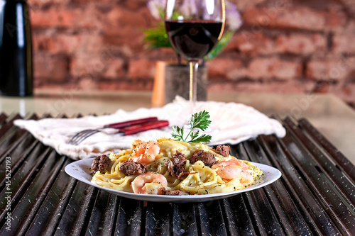 Spaghetti pasta with white sauce, meat, shrimp, mozzarella cheese and fresh basil on a white plate, red wine, on a black grill and rustic wall