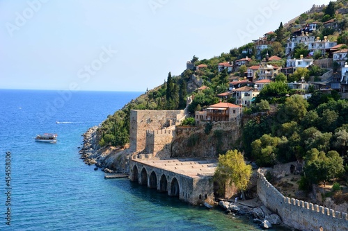 mountain with ancient fortress, green pine trees and cosy houses on bank of mediterranean sea with turquoise water and blue sky