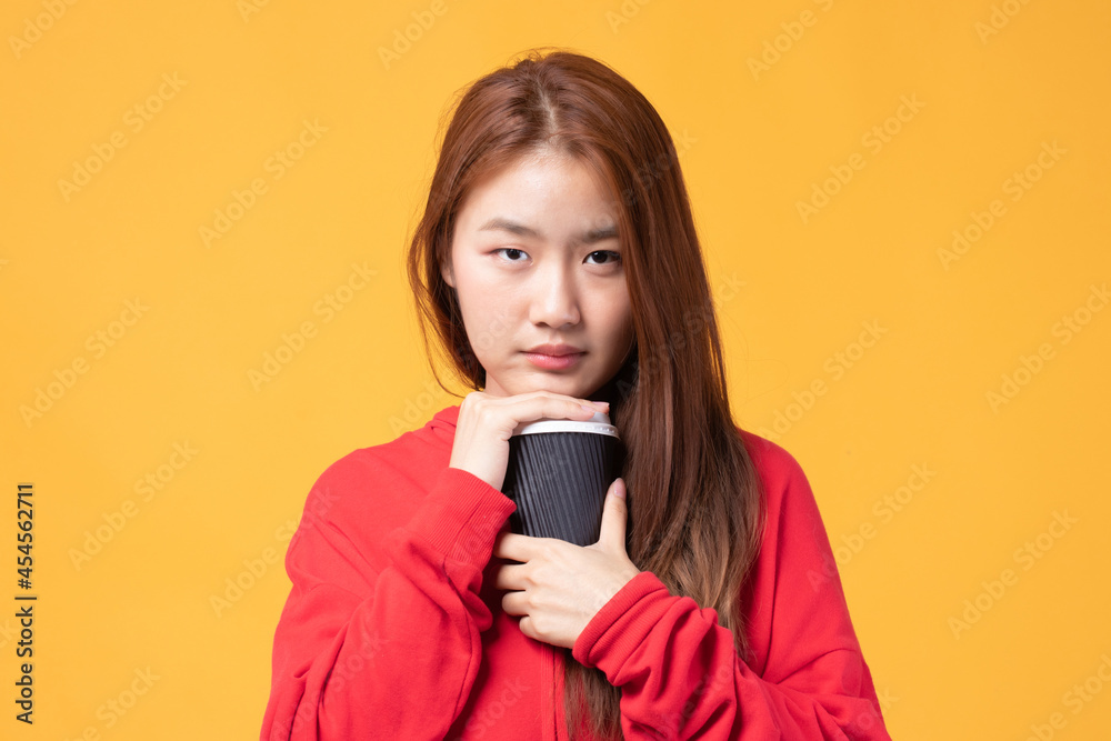 Portrait of a pretty young Asian woman smile and holding takeaway coffee cup wearing red sweater isolated on yellow background. Beautiful young girl enjoys coffee in the morning. Portrait of a pretty young Asian woman smile and holding takeaway coffee cup wearing red sweater isolated on yellow background. Beautiful young girl enjoys coffee in the morning.