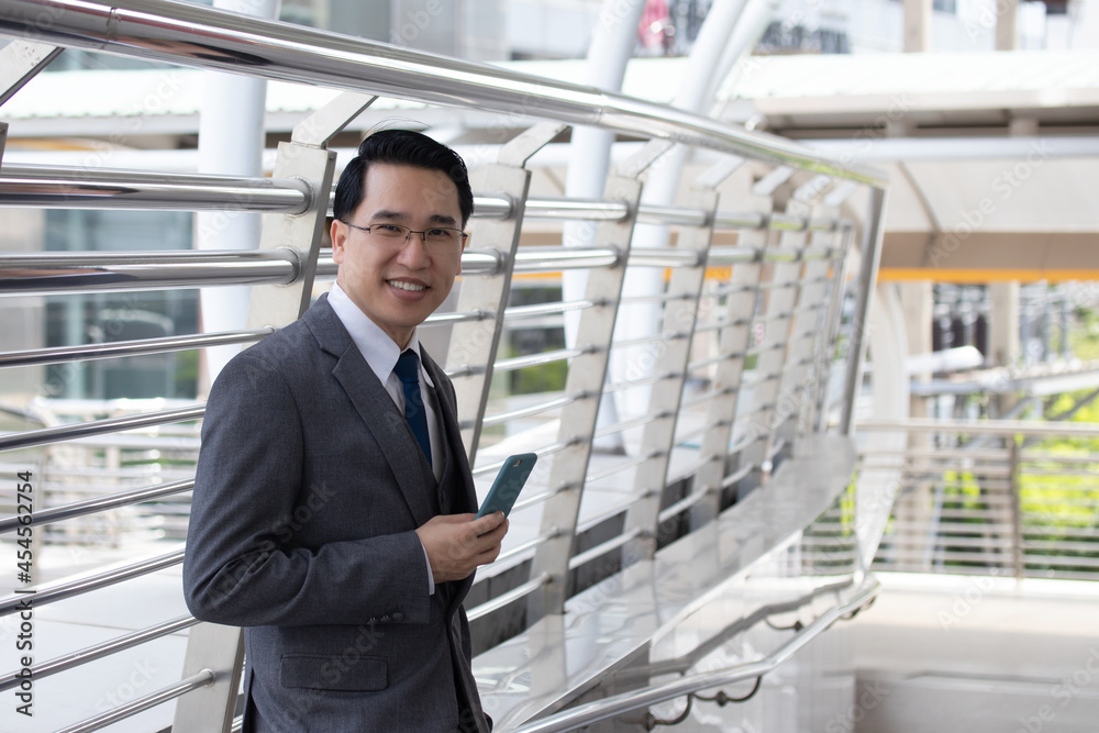 Asian businessman using mobile phone and smile outside of office in urban city. Asian man holding smartphone for business work. Asian businessman using mobile phone and smile outside of office in urban city. Asian man holding smartphone for business work.
