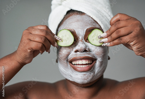 Young african woman with skin care cream holding fresh cucumber slices in front of her eyes - Beauty day and spa concept