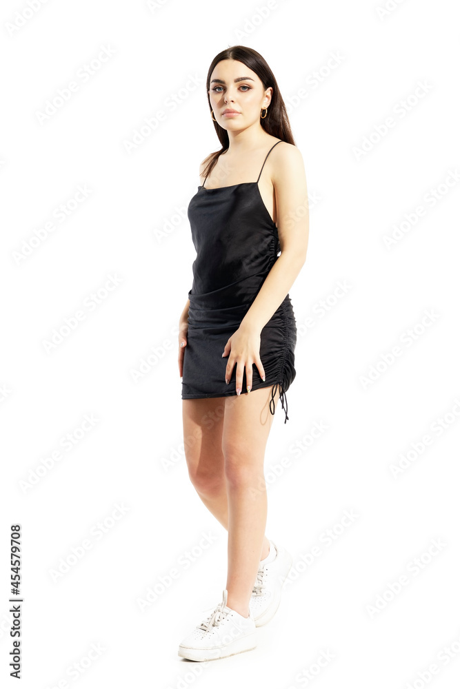 Fototapeta premium Stylish elegant young woman in short black satin dress walking and looking at camera. Full body portrait isolated on white background
