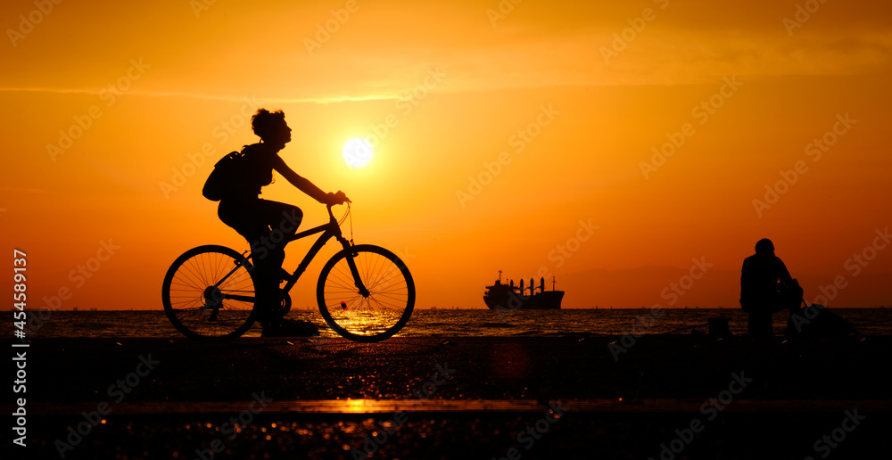 Obraz premium Silhouette of woman riding bicycle along the coastal road during the sunset.