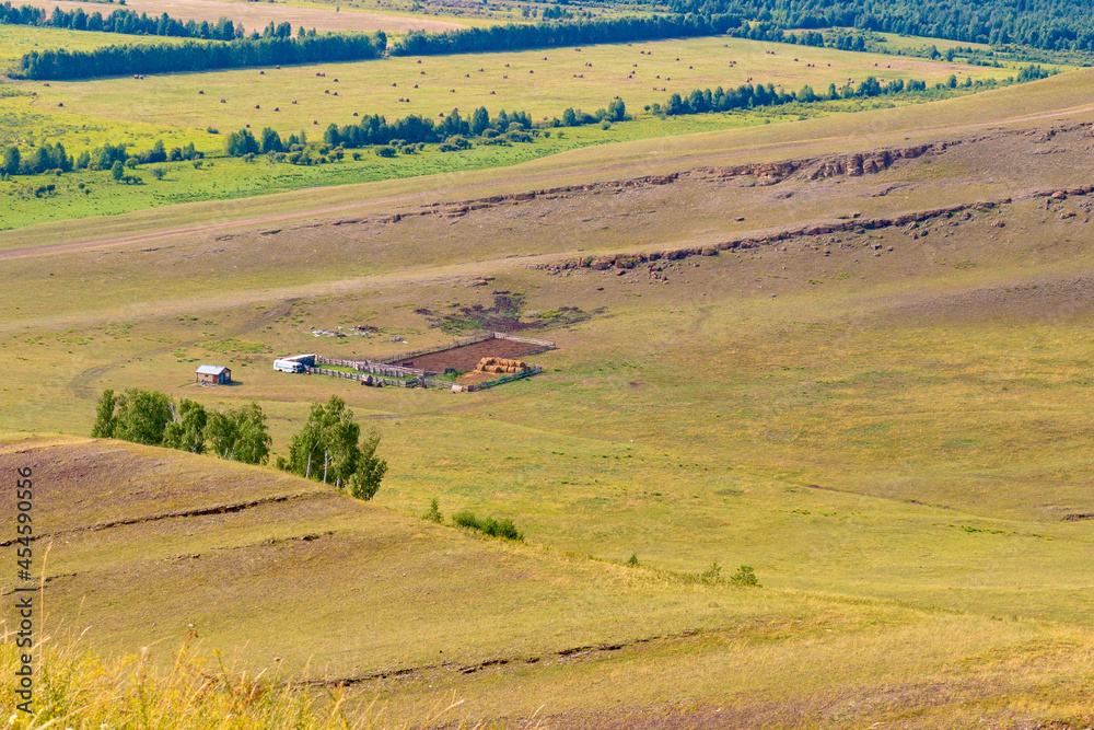 Aerial view of the pastures on green hillside of Khakassia on a summer day. Fenced cattle pen and small house in the distance in the meadow.