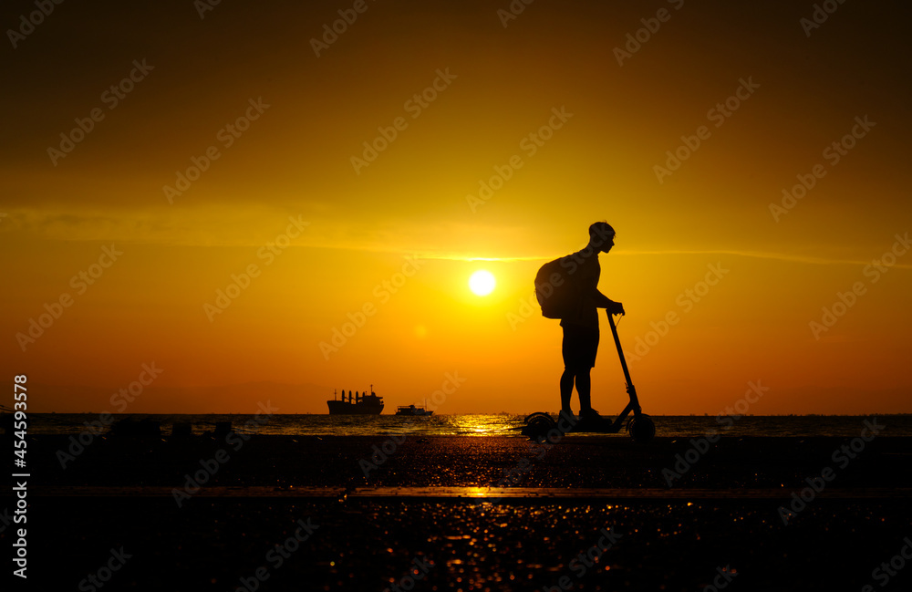 Silhouette of young man with backpack on an electric scooter on the embankment near sea at sunset