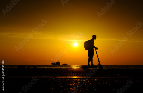 Silhouette of young man with backpack on an electric scooter on the embankment near sea at sunset © 22Imagesstudio