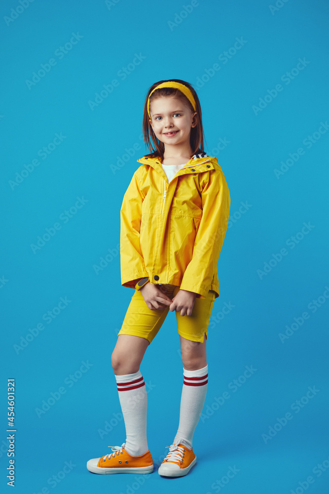 Full length excited little kid girl in yellow raincoat and sneakers smiling and keeping hands together against blue background Full length excited little kid girl in yellow raincoat and sneakers smiling and keeping hands together against blue background