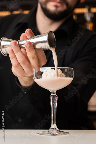 male bartender pouring to the measuring glass cup with ice cream an alcoholic drink from steel jigger on the bar counter on the blurred background