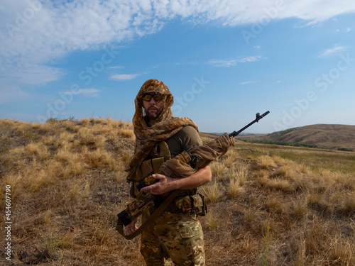 Sniper-mercenary in camouflage clothes under the scorching sun. He stands with a rifle and surveys the area.