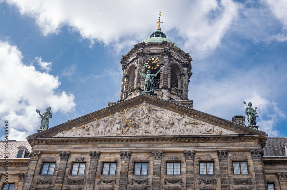 Amsterdam, Netherlands - August 14, 2021: Closeup of front brown stone monumental gable and clock tower of Royal Palace under blue cloudscape. Dense fresco and bronze statues.