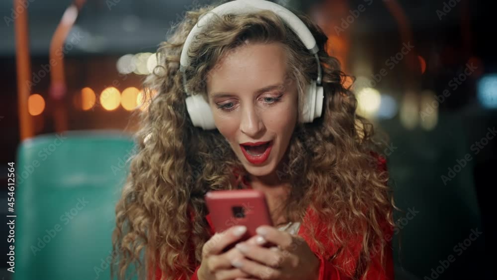 A curly woman is joying good message or news on smartphone in public transport. A girl got a positive answer or approval. Surprising and getting shocked. Concept of victory and gain. Night time. A curly woman is joying good message or news on smartphone in public transport. A girl got a positive answer or approval. Surprising and getting shocked. Concept of victory and gain. Night time.