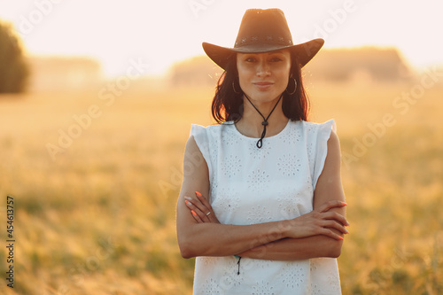 Woman farmer in cowboy hat at agricultural field on sunset with sun flare.