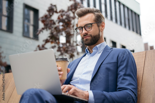 Young handsome bearded man wearing suit, stylish eyeglasses holding cup of coffee watching training courses. Confident businessman using laptop computer working online sitting at workplace 