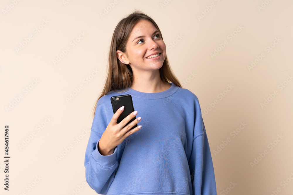 Young Lithuanian woman using mobile phone isolated on beige background looking up while smiling Young Lithuanian woman using mobile phone isolated on beige background looking up while smiling