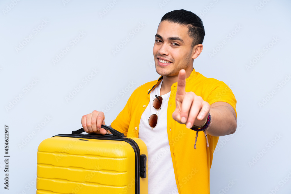 Asian handsome man isolated on blue background in vacation with travel suitcase and counting one Asian handsome man isolated on blue background in vacation with travel suitcase and counting one