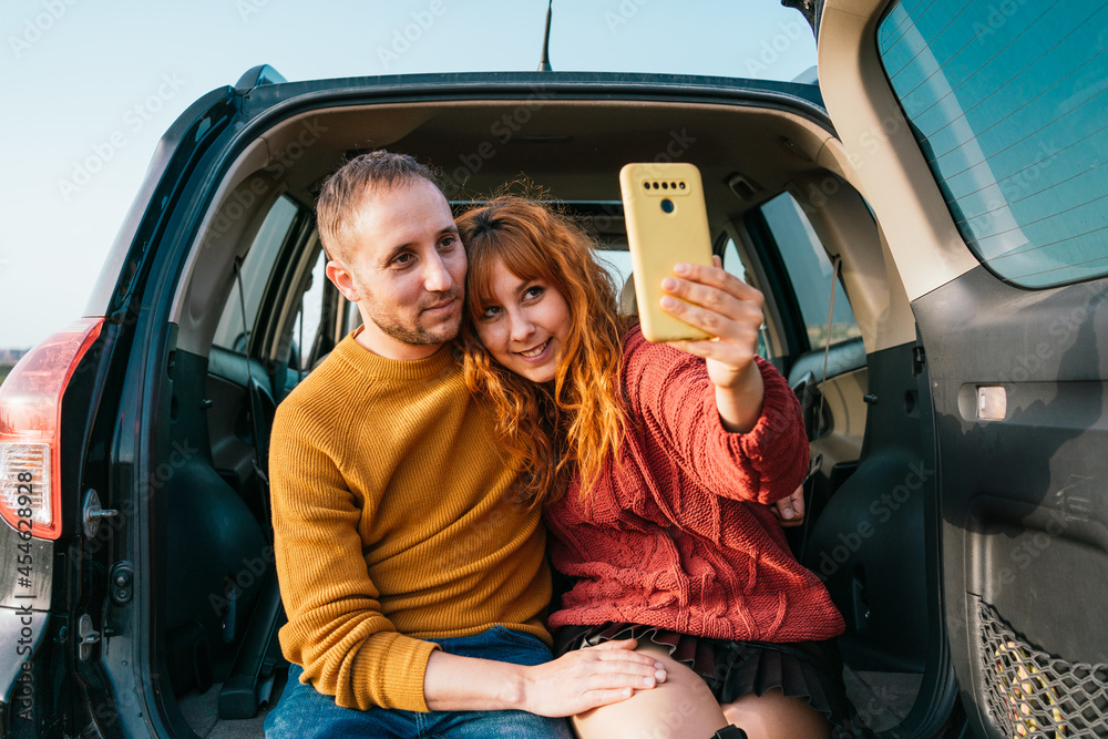 Young happy couple taking a selfie sitting on the car trunk