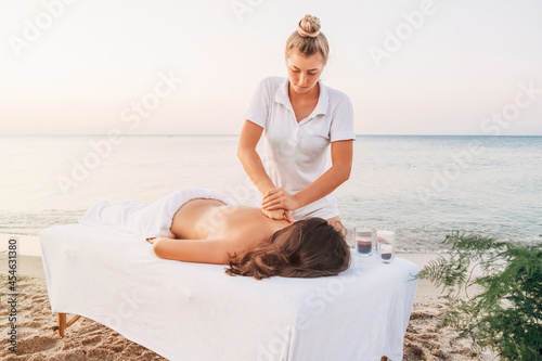 A young masseuse in white clothes makes a massage on the seashore at dawn.