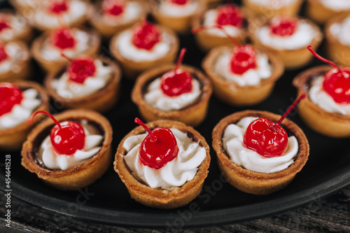 A lot of dough tartlets close-up with cream, cherries stand on a round tray, a plate on a wooden table. Banquet, buffet, photography