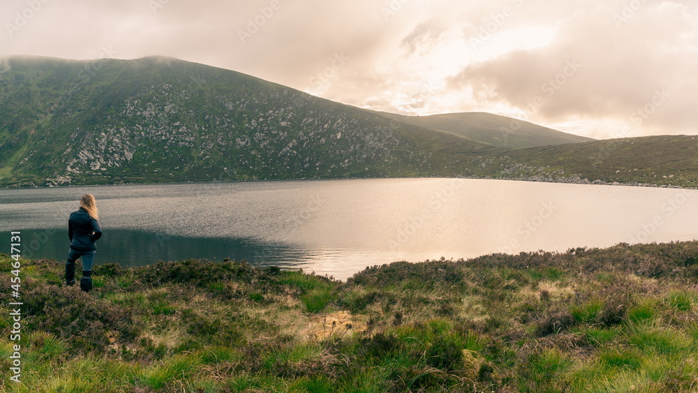 Happy backpacker. Young hipster woman standing, lake shore, beside a lake with epic, scenic mountain background. Wicklow, Ireland Happy backpacker. Young hipster woman standing, lake shore, beside a lake with epic, scenic mountain background. Wicklow, Ireland