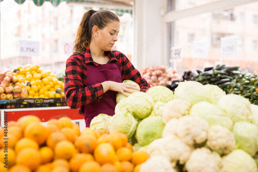 Young positive female seller displaying assortment of cabbages at grocery shop