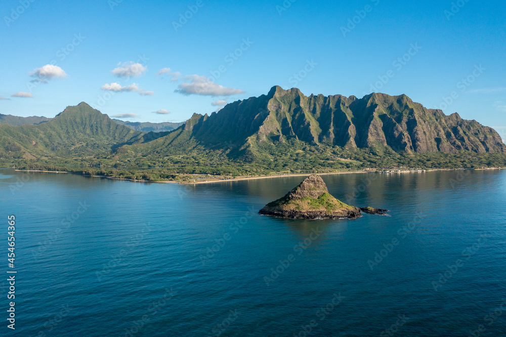 Naklejka premium Aerial view of Chinaman's Hat in Oahu, Hawaii, with the Ko'olau mountain range in the background. Morning golden light with a few puffy white clouds in a blue sky.