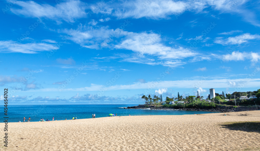 Daytime photo of Waimea Beach, Oahu, Hawaii. Blue sky with few white clouds, golden sand, church on right side of frame. Ample copy space in the sky.