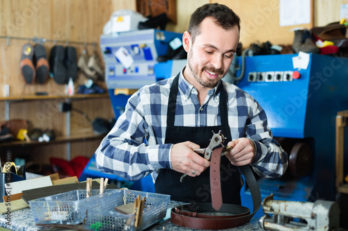 Young smiling glad friendly male worker making hole in belt in specialized workshop