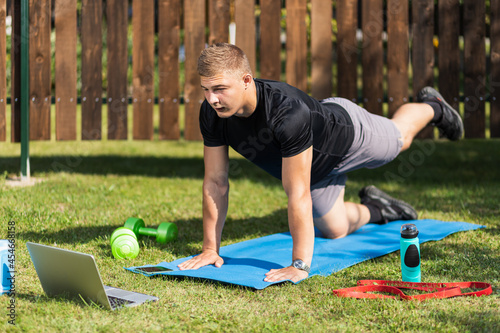 The young man goes in for sports outside. Cheerful sportsman with blonde hair makes the lunges on lawn in garden, next to there is a laptop with online training