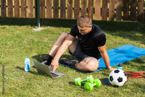 Close-up of a young man in a sports uniform is resting on lawn in garden, watches a movie and studies from a laptop , a social network. Student loafing around and putting off work and study