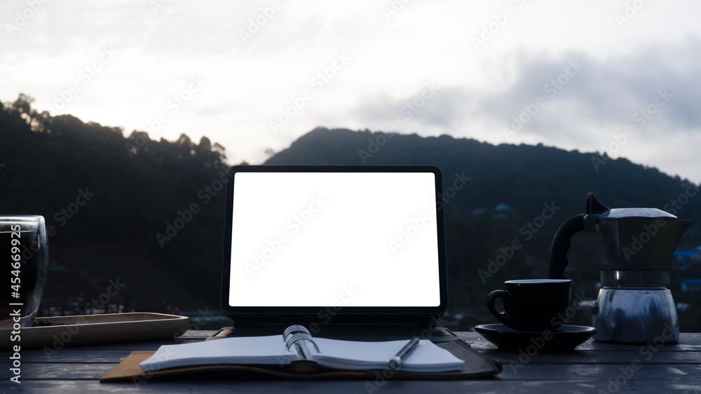 Fototapeta premium Computer tablet, notebook, moka pot and coffee cup on wooden table with mountain view in background.