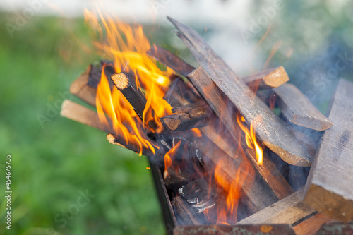 The firewood in the grill burns with a bright orange flame of fire on a natural green background. Preparation for cooking meat on the grill in nature. Fire flames and smoke 