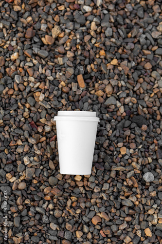 A white paper cup of coffee or tea lies on the rocky shore. A mug of hot drink on a pebble. Beautiful rocky background. free copy space.