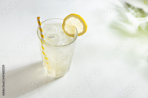 Lemon juice in soft light elegant white kitchen with green shadow of leaves in sunny day - lemonade in glass with straw, ice cubes, yellow lemon slice.