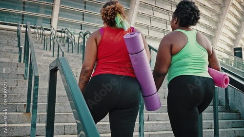 Two overweight african american women in sport clothes going upstairs to stadium, carrying fitness mats, tracking shot photo