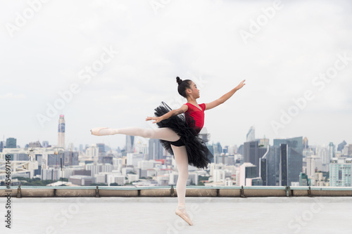 Asian ballerina ballet girl in red, black dress dancing on rooftop with cityscape view background. Young ballerina ballet girl dancing by stretching arms and leg on rooftop