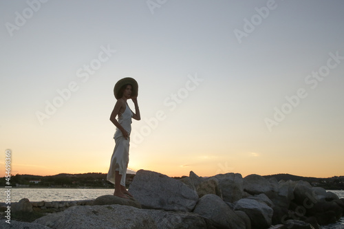 Young woman in long white dress and straw hat on the beach during sunset. Stylish summer vacation fashion.