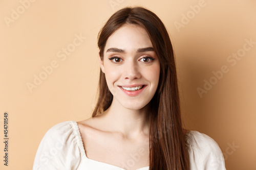 Close up portrait of happy woman with long hair and natural makeup, smiling cheerful at camera, standing on beige background